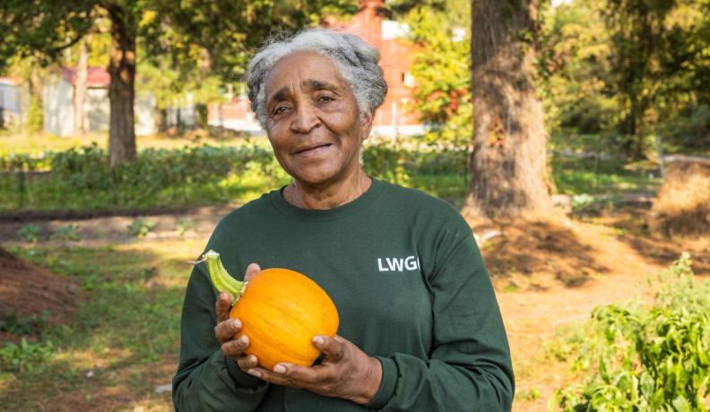 Cheryl Alston stands in a green long sleeve shirt holding a small pumpkin. She is looking calmly and pleasantly at the camera.