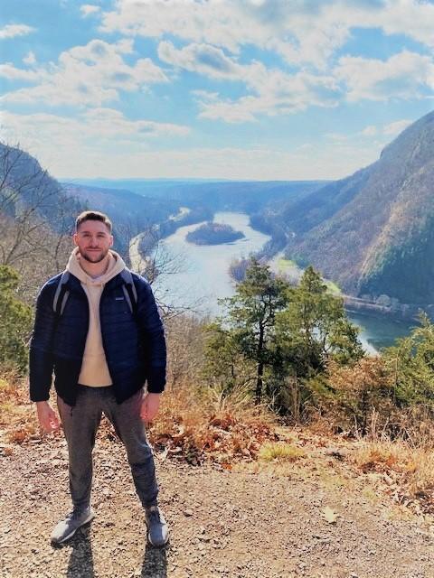 A man stands outside, a large river in a valley is in the background below him.
