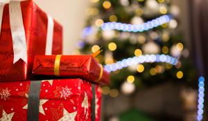 a stack of three presents wrapped in red, shiny paper are sitting in front a Christmas covered in yellow and blue lights.