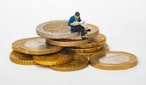 miniature figure of a person in a blue jacket sitting on top of a stack of gold coins.